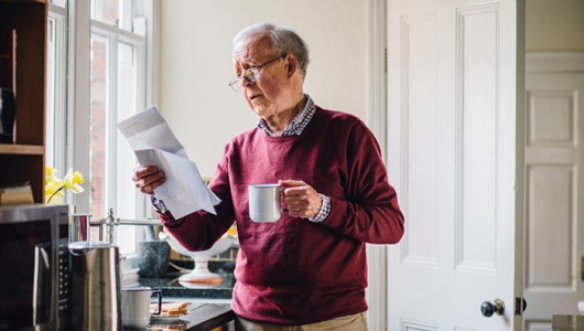 Senior man is standing in the kitchen of his home with bills in one hand and a cup of tea in the other. He has a worried expression on his face.